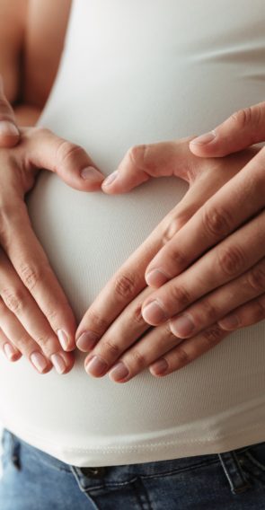 Close up portrait of man's and woman's hands making heart gesture over pregnant belly indoors